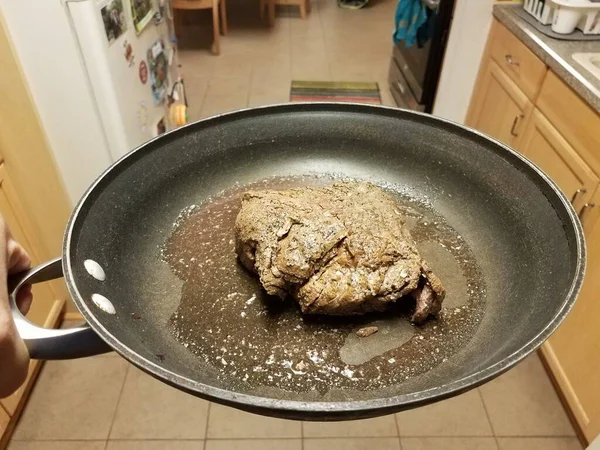 Mano sosteniendo la carne de res con jugos en sartén en la cocina —  Fotos de Stock