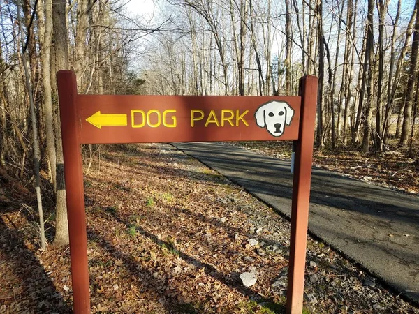 Wood dog park sign with yellow arrow and asphalt trail and trees — 스톡 사진