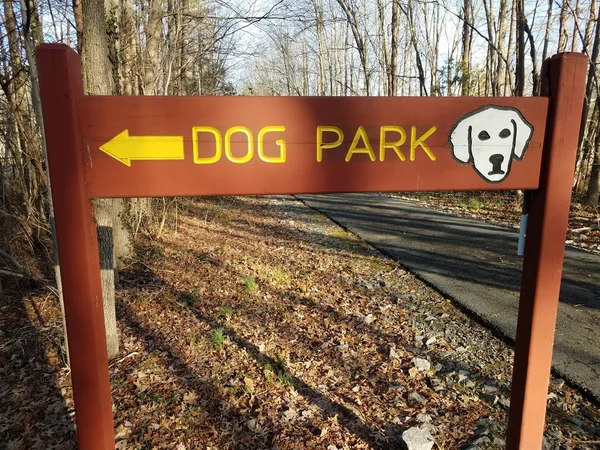 Wood dog park sign with yellow arrow and asphalt trail and trees — 스톡 사진