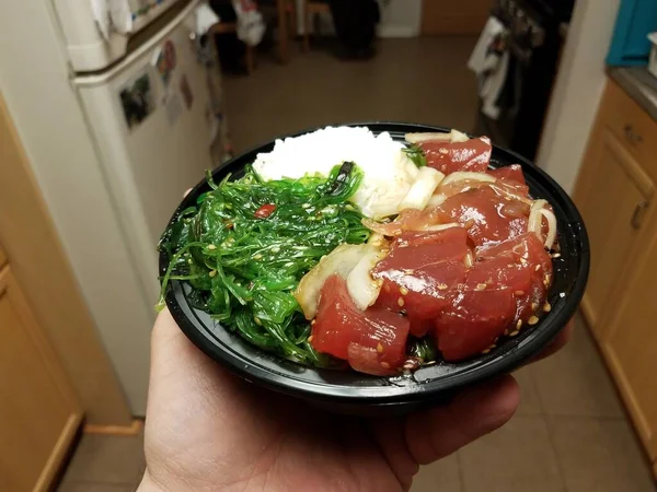 Hand holding bowl of raw tuna and green seaweed and rice in kitchen — 图库照片