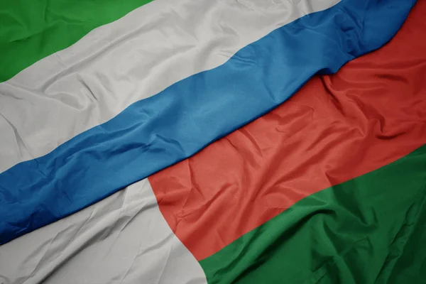 Ondeando colorida bandera de madagascar y bandera nacional de sierra leona . — Foto de Stock