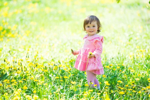 Una niña con un vestido rosa se ríe en un claro con dientes de león . —  Fotos de Stock