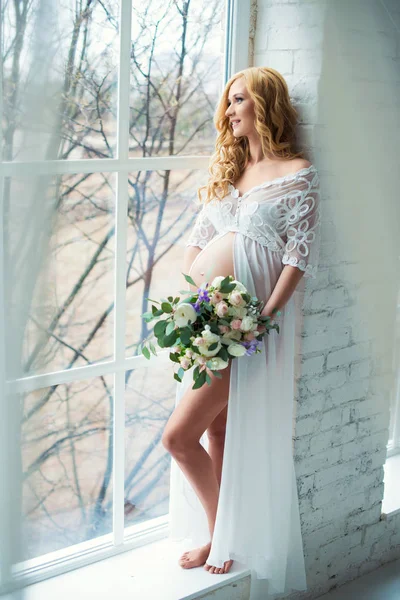Γοητευτική έγκυος κορίτσι κοιτάζει έξω από το παράθυρο με ένα μπουκέτο λουλούδια. — Φωτογραφία Αρχείου