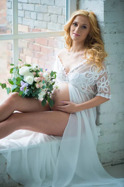 Mooi zwanger meisje glimlachend zittend op venster met boeket bloemen. — Stockfoto