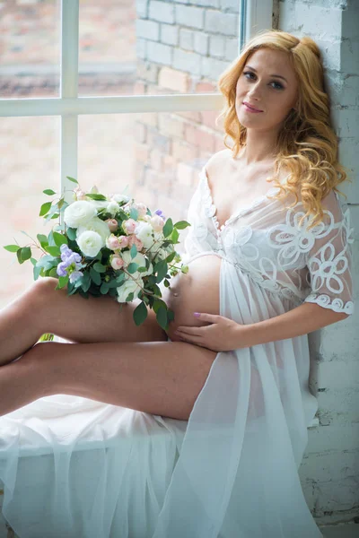 Menina grávida bonita sorrindo sentado na janela com buquê de flores . — Fotografia de Stock