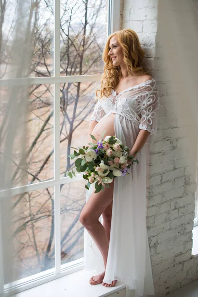 Encantadora menina grávida olha pela janela com um buquê de flores . — Fotografia de Stock