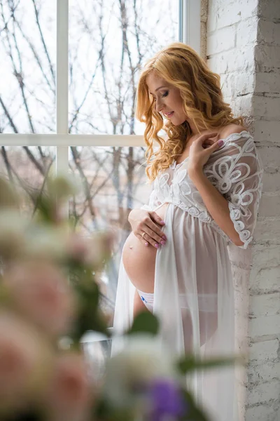 Γοητευτική έγκυος κορίτσι κοιτάζει έξω από το παράθυρο με ένα μπουκέτο λουλούδια. — Φωτογραφία Αρχείου