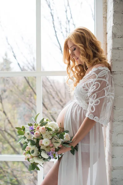 Charmante zwanger meisje kijkt uit het raam met een boeket van bloemen. — Stockfoto