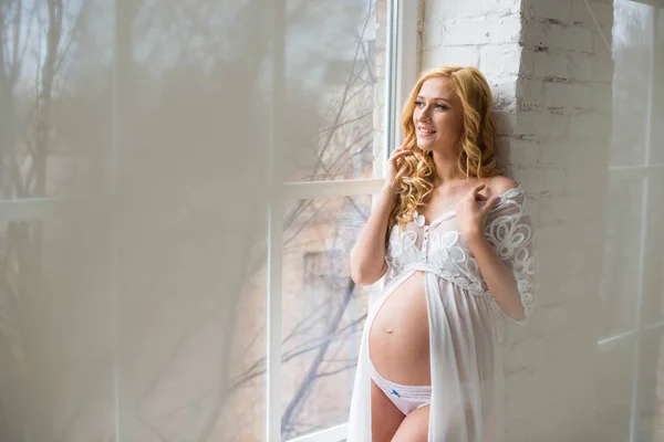Charmigt gravid flicka ser ut genom fönstret med en bukett blommor. — Stockfoto