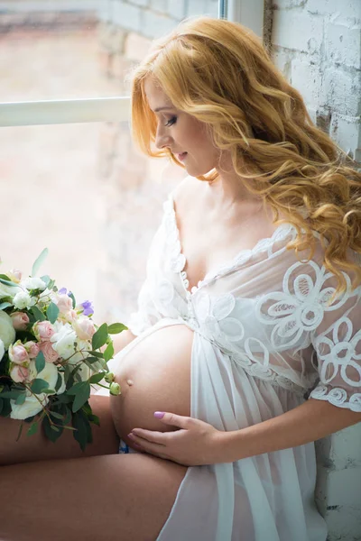 Mooi zwanger meisje glimlachend zittend op venster met boeket bloemen. — Stockfoto