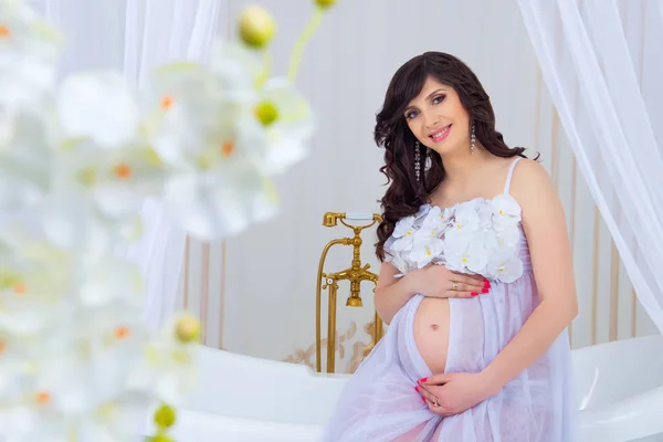 Jemný těhotenství. krásná těhotná ve světle bílých šatech s orchidejemi — Stock fotografie