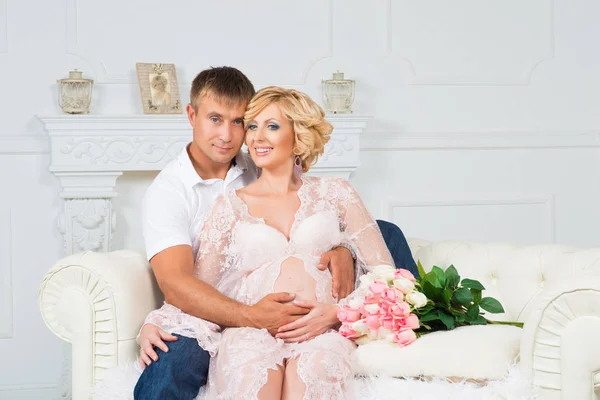 Feliz futuro padre con hermosa mujer embarazada linda sentado en el sofá con flores . —  Fotos de Stock