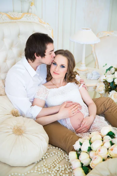 Casal bonito, mulher jovem grávida e homem abraçando com beijos de amor, sentado na cama, em um interior de casa . — Fotografia de Stock