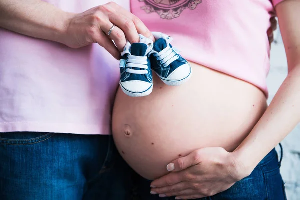 Детская обувь в руках молодого отца на фоне живота беременной женщины — стоковое фото
