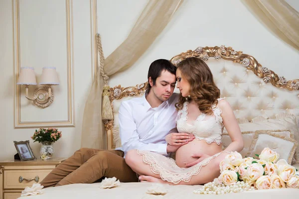 Hermosa pareja, mujer joven embarazada y hombre, abrazándose amorosamente sentado en la cama, en el interior de una casa . —  Fotos de Stock