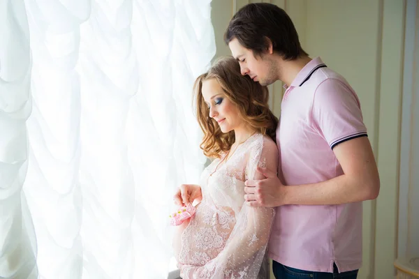 Botas de bebê nas mãos de uma futura mãe. Casal bonito, mulher jovem grávida e homem abraçando perto de uma janela . — Fotografia de Stock