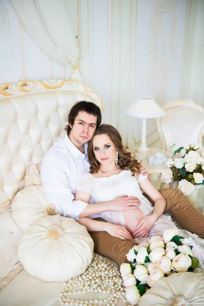Casal bonito, mulher jovem grávida e homem, abraçando amorosamente sentado na cama, em um interior de casa . — Fotografia de Stock