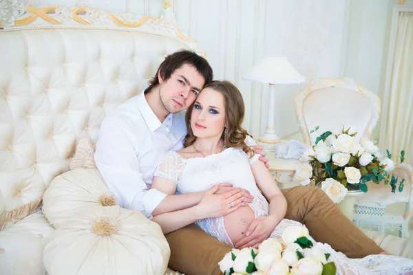 Prachtige paar, zwangere jonge vrouw en man, knuffelen liefdevol zittend op het bed, in een interieur. — Stockfoto