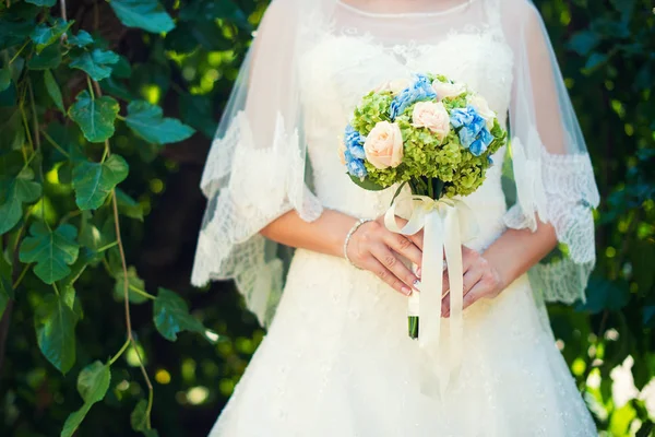 Noiva em um vestido de pé em um jardim verde e segurando um buquê de casamento de flores e vegetação. — Fotografia de Stock