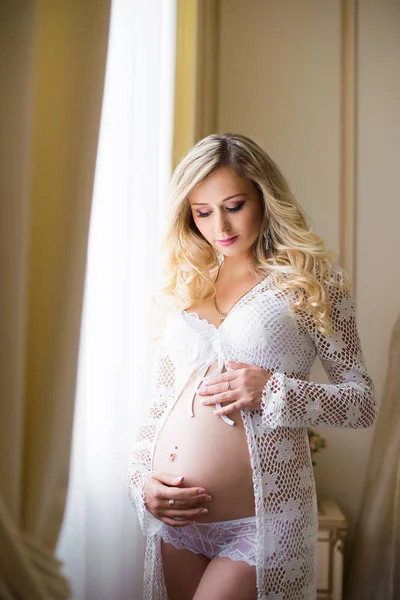 Charming pregnant girl is standing by the window looking at her tummy. Waiting for the baby. — Stock Photo, Image