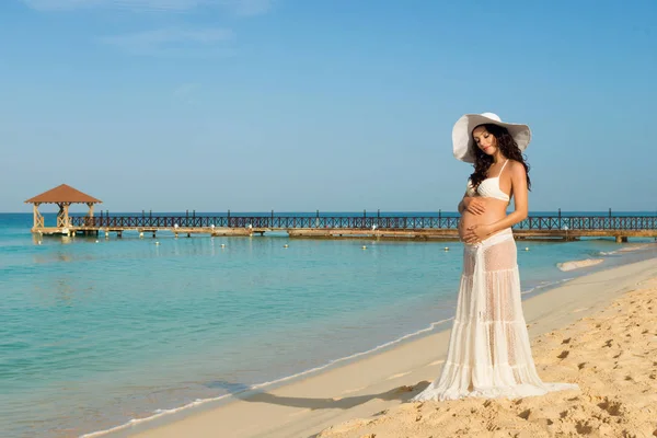 Beautiful pregnant woman on a sandy beach hugging her tummy. Dominican Republic, the Caribbean Sea. — Stock Photo, Image