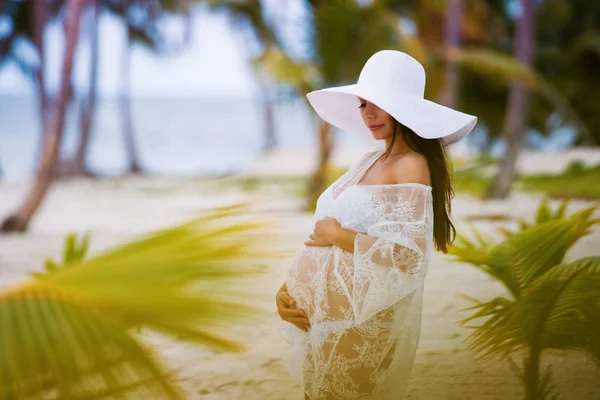 Bella ragazza incinta in abito bianco e cappello a tesa larga sulla spiaggia vicino alle palme — Foto Stock