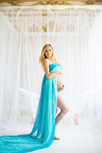 Красивая беременная женщина. Привлекательная блондинка дотрагивается до голого живота, позируя в обдувном платье из драпировки, летящем на ветру. — стоковое фото