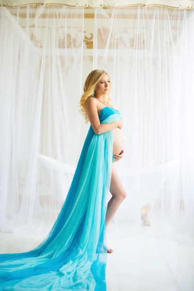 Красивая беременная женщина. Привлекательная блондинка дотрагивается до голого живота, позируя в обдувном платье из драпировки, летящем на ветру. — стоковое фото