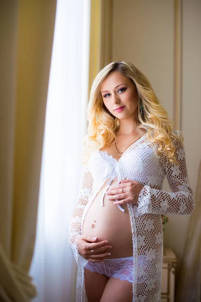 Charmigt gravid flicka står vid fönstret tittar på sin mage. Väntar på barnet. — Stockfoto