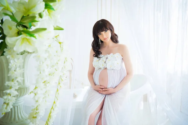 Suave expectativa de maternidad. Hermosa embarazada en vestido blanco claro con orquídeas — Foto de Stock