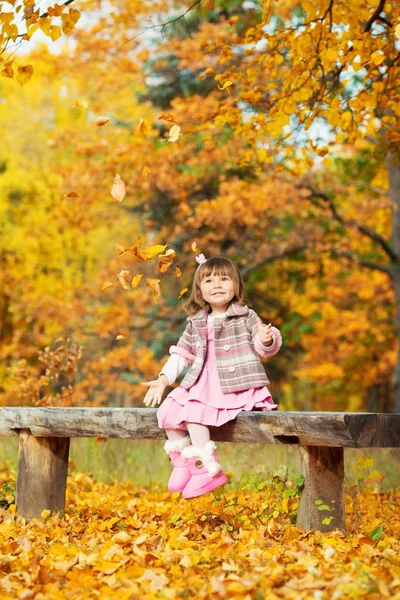 Niña feliz sentada en un banco riendo y jugando con hojas. En la naturaleza, caminar al aire libre —  Fotos de Stock