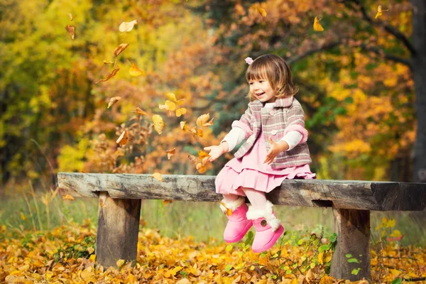 Счастливая маленькая девочка, сидящая на скамейке и смеющаяся и играющая с листьями. На природе гулять на свежем воздухе — стоковое фото