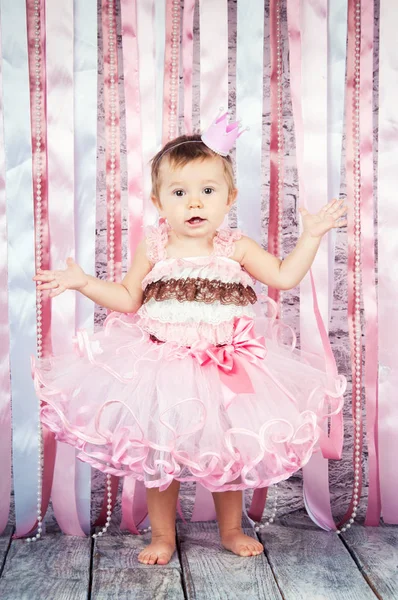 A sweet little girl in a crown and a beautiful pink dress is raising her hands on the stage, a background of pink ribbons — Stock Photo, Image