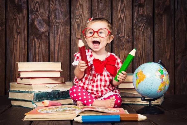 Happy a nice little girl with glasses and pencils against the background of books and a globe — Stock Photo, Image