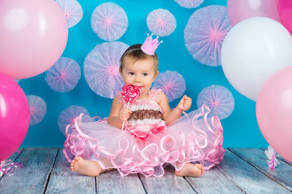 Lustiges Kind mit einem Lutscher in Herzform, glückliches kleines Mädchen mit Krone auf dem Kopf, das einen großen Zuckerlutscher isst, isoliert auf hellblauem Hintergrund mit rosa Kugeln, Studio. — Stockfoto