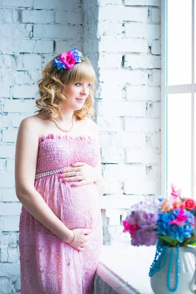 Schwangere lächelt, blickt auf Bauch, hält Blumen in der Hand. rosa Kleid. — Stockfoto