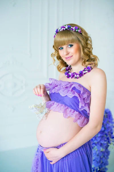 Šťastné těhotné ženy v fialová dlouhá sukně představuje v ateliéru na pozadí květů — Stock fotografie