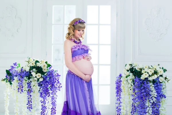 Eine glückliche schwangere Frau in lila langem Rock posiert im Studio vor einem Hintergrund aus Blumen — Stockfoto