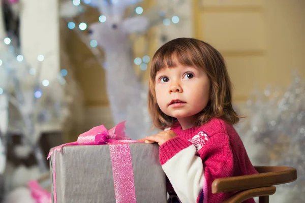 Een klein meisje met een cadeau in haar handen zit in een slee op de achtergrond van een hert. — Stockfoto