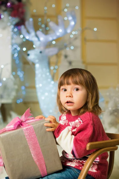 Een klein meisje met een cadeau in haar handen zit in een slee op de achtergrond van een hert. — Stockfoto