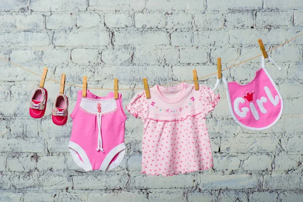 Ein rosa Kinderleibchen, ein Lätzchen, ein Kleid und rote Schuhe für ein Mädchen, trocken auf einem Seil an einer weißen Ziegelwand. — Stockfoto
