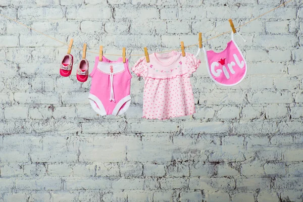 Um bodik rosa das crianças, um babador, um vestido e sapatos vermelhos para uma menina, seco em uma corda contra uma parede de tijolo branco . — Fotografia de Stock