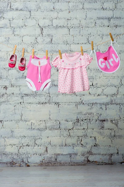 Дитячий рожевий босик, нагрудник, сукня і червоне взуття для дівчинки, сухе на мотузці на білій цегляній стіні . — стокове фото