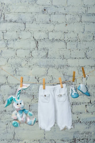 Childrens white bodik, boots, panties and a toy rabbit dry on a rope against a white brick wall. — Stock Photo, Image