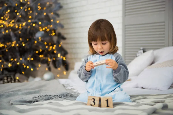 Small cute little girl is 2 years old sitting near Christmas tree and looking at the calendar. 31th of December. — Stock Photo, Image