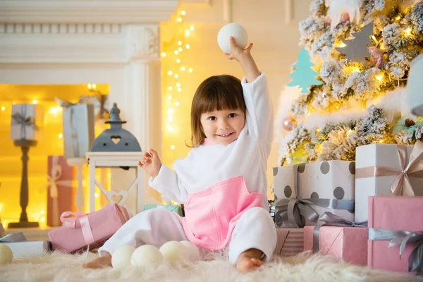Petite fille souriante émotionnelle assise en pyjama avec des cadeaux de Noël près de l'arbre du Nouvel An et jouant aux boules de neige — Photo
