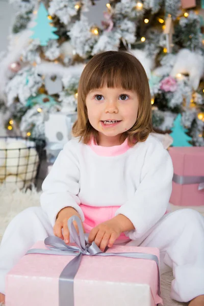 Niña divertida en pijama de lana blanca y rosa sentada cerca del árbol de Navidad y desempacando el regalo de Año Nuevo —  Fotos de Stock