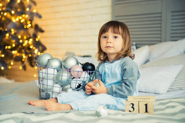 Small cute little girl is 2 years old sitting near Christmas tree and looking at the calendar. 31th of December. — Stock Photo, Image