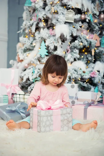 Θετική χαρούμενο μωρό κορίτσι που κάθεται με το δώρο Χριστουγέννων κοντά στο χριστουγεννιάτικο δέντρο. Ευτυχισμένος ο καινούριος χρόνος — Φωτογραφία Αρχείου