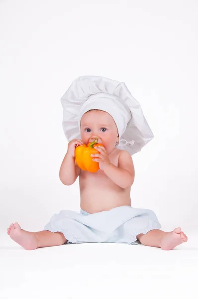 A funny little girl in a chefs clothes is eating pepper. — Stock Photo, Image